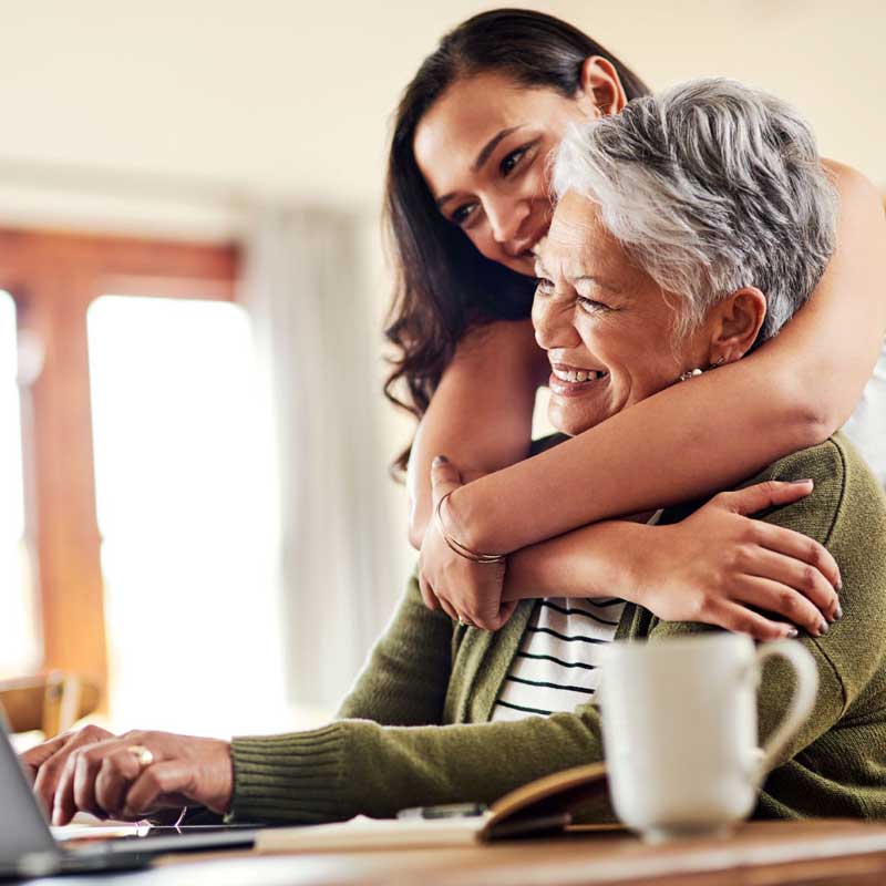A young woman embraces her smiling grandmother as they share a joyful moment together, seated at a table with a laptop.