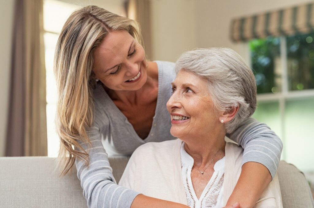 A senior woman sitting on a sofa is hugged from behind by her adult daughter.