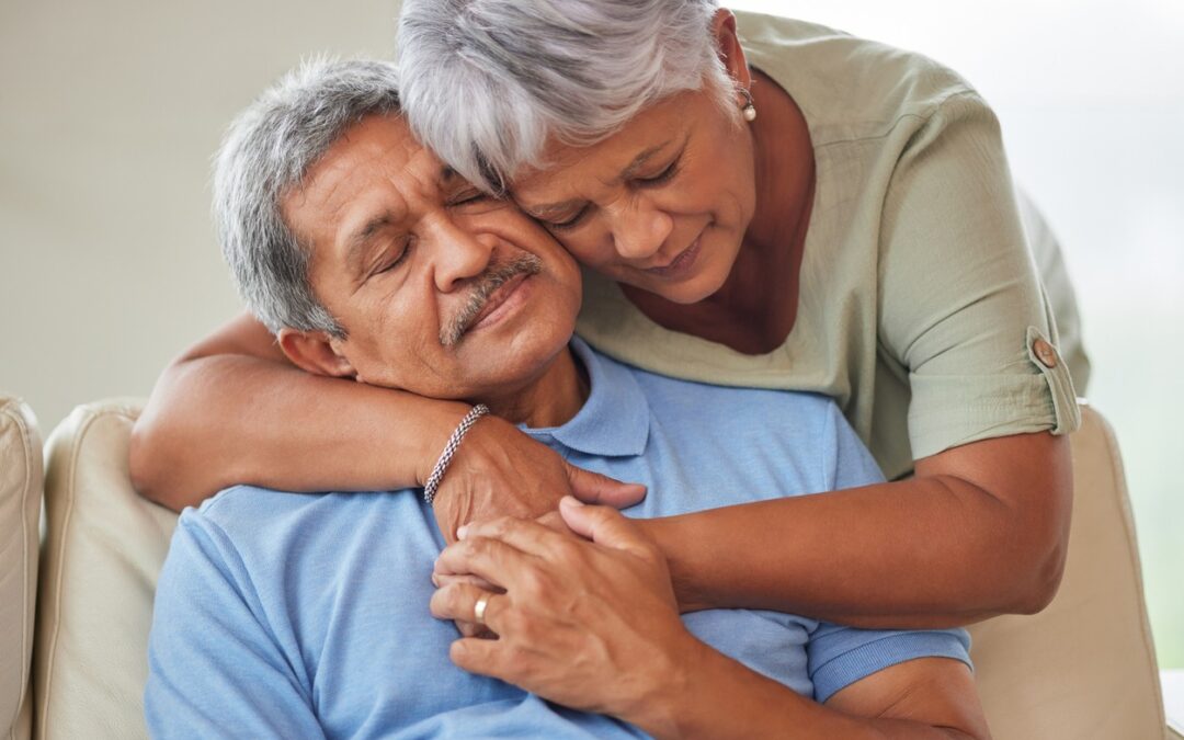 An elderly Hispanic woman embraces her Hispanic husband while he's sitting.