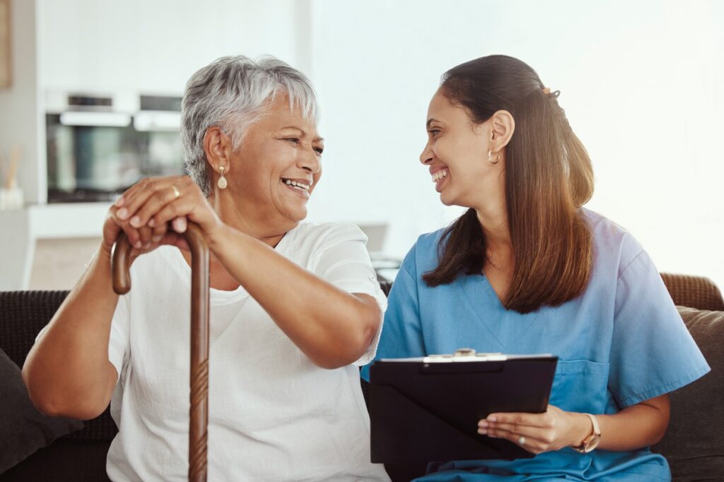 A palliative care nurse with an elderly woman holding a cane in her home.